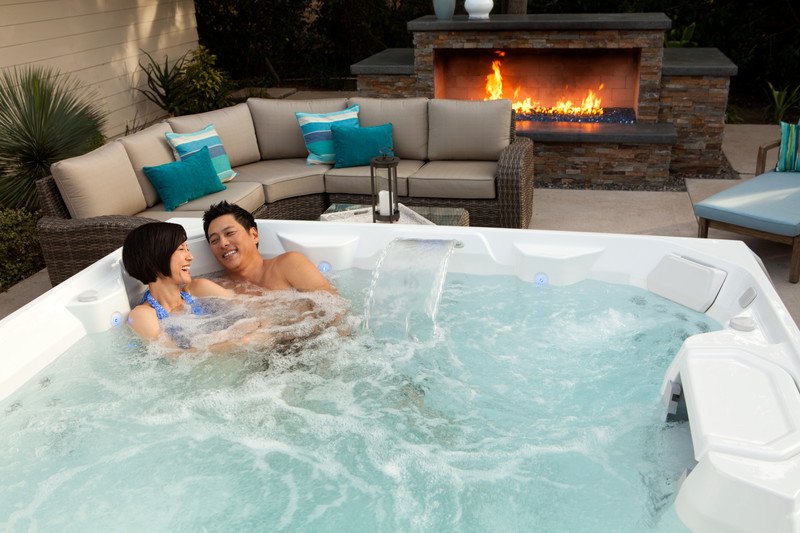 A blissful couple relaxes in a bubbly hot spring spa hot tub with a water cascade and firepit in the background. Chlorine keeps the water clean, adding to the serene experience.