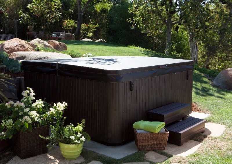 A brown hot tub with a closed cover, surrounded by greenery and rocks. A basket with towels and a step stool are placed near the tub.