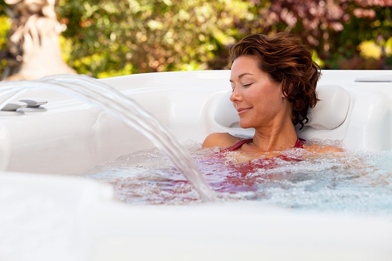 A woman is peacefully relaxing in a hot tub, with her eyes closed and water cascading gently around her.